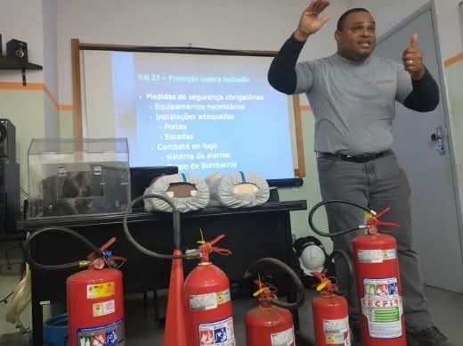 Instrutor ministrando treinamento de NR 23 (Prevenção contra Incêndio) com diversos extintores展示dos e tela de apresentação ao fundo.